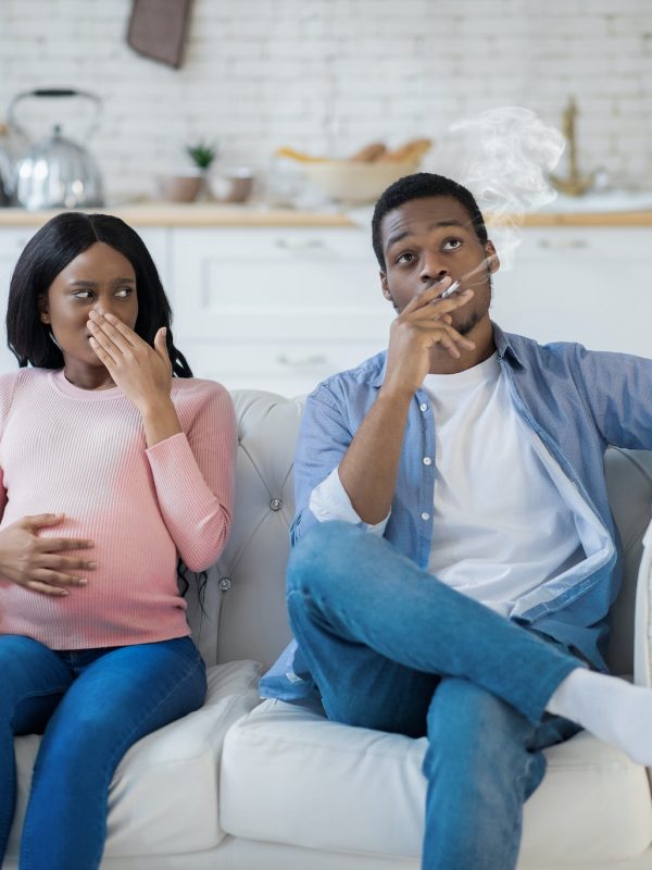 Nicotine abuse during pregnancy concept. Selfish black man smoking near his expectant wife at home
